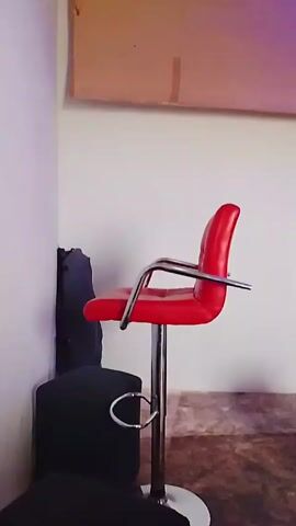 The video shows a small, sparsely furnished room. In the foreground, there is a red bar stool with a metal base. Behind it is a dark object, possibly another chair. The walls are white, and the floor is partially visible and appears to be textured. On the wall, there is a rolled-up beige window shade. The ceiling is white with a grid pattern and recessed lighting, one of which emits a blue light. A purple hue is present, possibly due to the lighting.