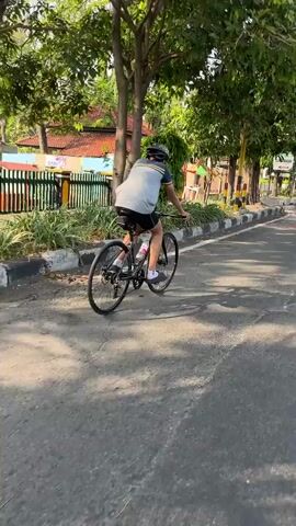 A person wearing a helmet, t-shirt, and shorts is riding a bicycle on an asphalt road. The person is wearing a grey t-shirt with a blue and yellow stripe on the collar and sleeves. The person is also wearing black shorts and white shoes. The bicycle is black and has thin tires. The person is wearing a helmet that is black and grey. The road is grey and has white lines on the sides. There are trees and green foliage on the left side of the road. In the background, there is a fence and a building with a red roof. The sun is shining, and there are shadows on the road. The person is looking forward and appears to be pedaling the bicycle.
