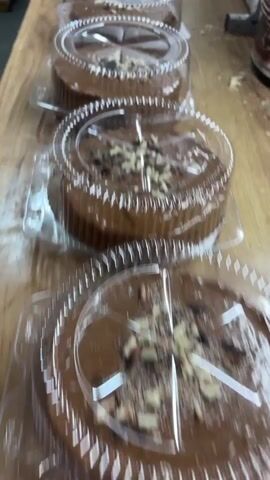 The video captures a close-up view of multiple small, round chocolate cakes, each meticulously packaged in individual clear plastic containers with domed lids. The cakes are uniformly decorated with what appears to be a scattering of light and dark chocolate shavings or chopped nuts on top of a smooth chocolate glaze. They are arranged in a straight line on a light-colored wooden surface, suggesting a display or preparation area in a commercial setting like a bakery or kitchen. In the background, part of a large white bucket or container is visible, bearing text that includes "6 kg." and some Cyrillic letters, possibly indicating an ingredient like chocolate. The camera pans slowly along the row of cakes, showcasing their consistent presentation. No people are visible, but the scene implies activity related to food preparation and packaging for sale.