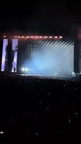 The video shows a crowded concert venue at night. A large stage is illuminated with bright lights and smoke. Silhouettes of performers can be seen on stage. The audience is mostly dark, but many people are holding up their phones to record the show. The overall atmosphere is energetic and exciting.