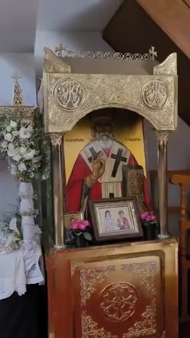 A video pans across an ornate religious altar. On the right, a large golden icon depicts a saint in red and white vestments, holding a book and a cross. Below it are smaller icons and pink flowers. To the left, a more elaborate floral arrangement covers a shrine, featuring a crown-like structure adorned with white and pink flowers, and a draped white cloth. The background appears to be the interior of a church, with a speaker mounted on the wall.