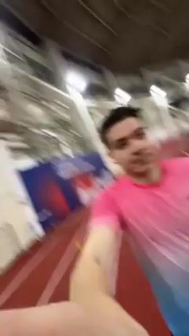 A man is recording himself with his outstretched arm while running on an indoor track. He is wearing a pink and blue ombré T-shirt, black shorts, and black shoes, and he has wireless earbuds in. The track is red with white lines. In the background, there is a blurry view of the indoor athletic facility. The lighting is bright.
