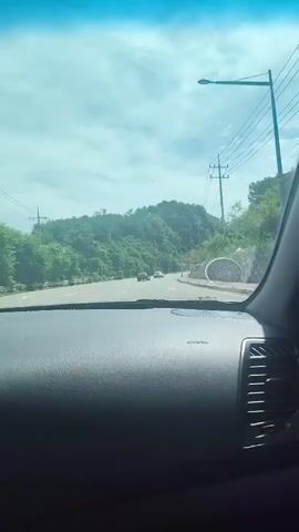 The video shows a view from inside a car as it drives along a road. The dashboard is visible in the foreground. The road is surrounded by lush green hills and trees. There are a few other cars on the road ahead. Overhead, power lines and a street light are visible against a bright blue sky with some clouds. The car is moving forward at a steady pace. The side mirror also shows a glimpse of the passing scenery, which includes a wall made of concrete blocks and some fencing. There appears to be a small white tent or canopy in the distance on the right side of the road.