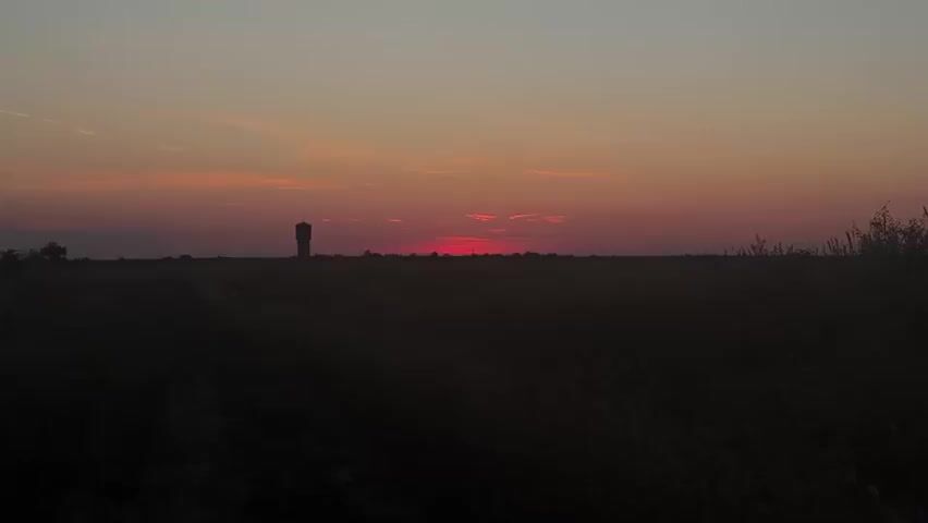 A wide shot captures a rural landscape at dusk. The sky transitions from a pale blue at the top to a gradient of orange and deep red near the horizon, with thin clouds streaking across. A silhouetted water tower stands in the distance on the left, its dark form stark against the vibrant sky. The foreground is dominated by dark, indistinct shapes of tall grass and vegetation, obscuring the ground and creating a silhouette effect. The overall atmosphere is tranquil and serene, with the setting sun casting a warm glow on the scene.
