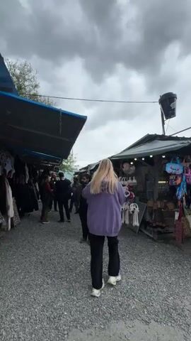 The camera follows a woman with long blonde hair wearing a purple sweatshirt as she walks through an outdoor market on a cloudy day. The ground is covered in small gray stones. People are walking in both directions and standing at the stalls, which are covered by tarps. The stalls are selling various items, including clothing, jewelry, and souvenirs.