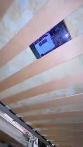 A mobile phone is wedged between the wooden slats of a bed frame, playing a video. The video on the phone features a purple and black background with text and images. The camera focuses on the phone's screen and then pans out to show the underside of the bed, including the frame and mattress. A glimpse of the room is visible through a gap under the bed, revealing a door and a patterned curtain or blanket. The camera zooms in and out, showcasing the phone's screen and the surrounding bed structure. A male voice is heard in the background, speaking in Spanish.