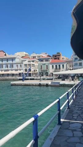 The video pans across a scenic waterfront view in Agios Nikolaos, Greece. The scene is a picturesque harbor filled with clear, turquoise water. Buildings line the waterfront, showcasing a mix of architectural styles with pastel and white facades, balconies, and awnings. Some of these buildings appear to be hotels or restaurants. Several boats are docked in the harbor, including a prominent red and white ferry. The sky is a vibrant, clear blue, indicating a sunny day with excellent weather. The sun is high, suggesting it is midday. A paved walkway with a white and blue railing runs along the edge of the water in the foreground, implying a public promenade or harbor front. There are a few people visible walking along the waterfront and seated at outdoor cafes, suggesting a leisurely atmosphere. In the distance, a hill is visible with some structures on it. No text is clearly visible in the video. The overall impression is a peaceful and beautiful Mediterranean coastal town.