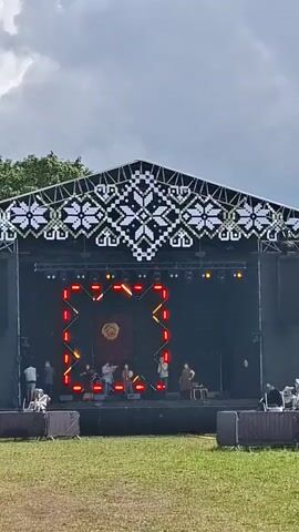 A wide shot captures a festival stage setup outdoors. The stage is covered by a roof with a decorative, white and black pixelated pattern along its edge. A large LED screen displays a pulsing red square with a circular floral design in the center. A group of people, likely performers, are on the stage. Some are holding instruments, suggesting a musical performance. In front of the stage, rows of empty seating or barriers are visible. The ground in the foreground is covered in green grass. The sky above is overcast with dramatic clouds.