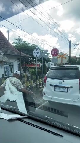 The video shows a view from inside a vehicle stopped at a railroad crossing. A white Toyota Avanza is in front of the filming vehicle, with its license plate P 1847 WA visible. To the left of the Avanza is a man sitting on a motorcycle, seemingly using his phone. Various traffic signs are present, including a height restriction sign (5.5m), a STOP sign, and the standard railroad crossing symbol. A passenger train is passing in the background. Other motorcyclists are also waiting at the crossing. The sky is partly cloudy, and utility lines are visible overhead. The environment appears to be a typical urban or suburban area.