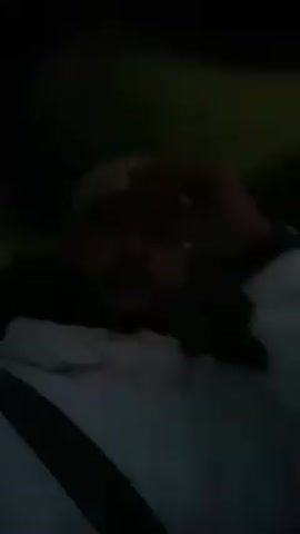 A person wearing a white hoodie and a headband is sitting in a car, talking to the camera. The scene is dark. The person is wearing a seatbelt.