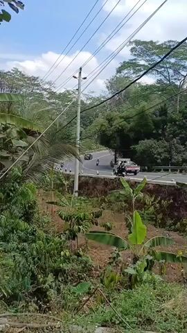 A highway curves through a lush, green, tropical environment. Telephone poles and overhead wires are visible. The road has a guardrail and a stone retaining wall on the side. Vegetation, including banana trees, grows on a slope beside the road. Vehicles, including a car, motorcycles, and a small truck, are seen traveling on the road. The sky is blue with some clouds.
