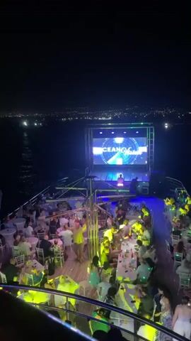This is a nighttime aerial shot of a party on a boat. The boat is crowded with people sitting at tables and standing. There is a large screen at the front of the boat displaying visuals and a DJ booth with a person visible. Colorful lights, primarily green and blue, illuminate the scene. In the background, a distant city skyline is visible, with lights reflecting on the dark water. The camera is panning slowly to the right.