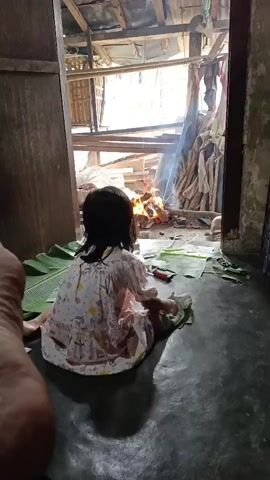 The media shows a young girl sitting on the floor in a room with a dirt floor and a partially open doorway. She is wearing a light-colored dress with a floral pattern. In front of her are several large green leaves, and she is holding a white cloth with green stripes. She appears to be interacting with something on the ground, possibly drawing or writing on paper, as a red pencil or crayon is visible.

Through the doorway, a fire is burning, with smoke rising. The background outside the doorway suggests a rustic outdoor setting with wooden structures and debris. The lighting indicates it is daytime, possibly mid-morning or afternoon, with sunlight filtering into the room. The overall impression is of a simple, perhaps rural, domestic scene.

The media's metadata specifies the location as Bunut, Indonesia. No text is visible within the media itself. The mood conveyed is one of quiet activity, with the child focused on her task amidst the backdrop of a fire and the surrounding environment.