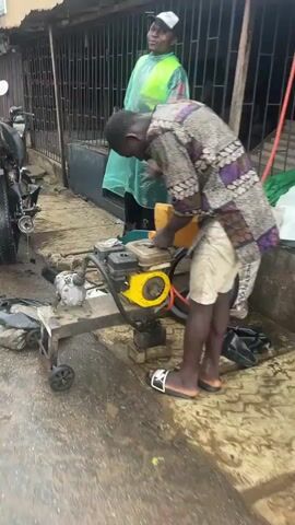 In an outdoor setting, a man wearing a patterned shirt and light-colored shorts bends over a piece of machinery that appears to be a small engine mounted on a wheeled frame. He is wearing slide sandals. Beside him is a yellow jerrycan. The ground is wet, and puddles are visible. To the left, part of a motorcycle is visible. Behind the man, another individual stands, wearing a black cap and a transparent green raincoat. They are standing near a fenced area. A black bucket is on the ground nearby. The focus is on the first man's interaction with the machinery, suggesting he is either repairing or operating it.