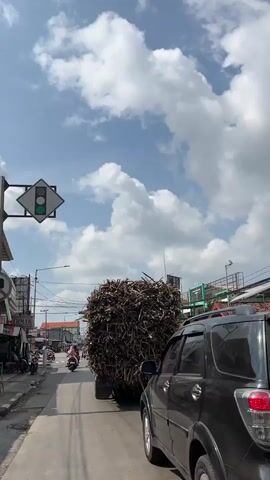 The video shows a view from a vehicle on a street, likely in a town or city, with a clear blue sky and fluffy white clouds above. A large truck heavily loaded with what appears to be dried sugarcane stalks or similar plant material is moving ahead. The truck is a yellow flatbed with blue trim. To the left of the truck, there are buildings with shops and storefronts, as well as power lines and a traffic light. Several motorcycles and a silver van are also visible on the road, suggesting moderate traffic. A black car is partially visible in the foreground on the right side of the frame. The overall impression is of a sunny day in a busy, possibly tropical, urban environment.