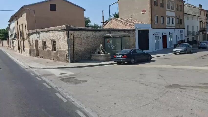 A black car is parked on a street corner next to a weathered stone building. In front of the building is a fountain shaped like a lion. A bus stop shelter is located behind the fountain. The street is paved and there are sidewalks on either side. Buildings line the street in the background. The sky is clear and blue. The video is filmed from a moving vehicle.