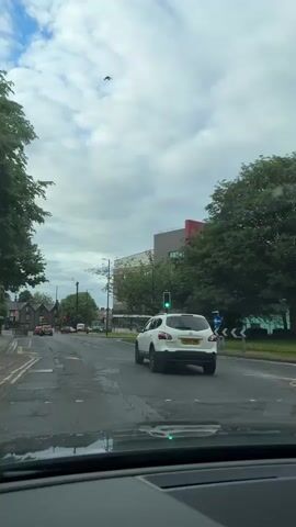 From the perspective of inside a vehicle, the video shows a view of a street with cars and buildings on a cloudy day. The traffic light is green. A white SUV with a license plate YH63 YNE moves forward through the intersection. The buildings visible are a mix of residential houses and a larger, modern building with a red and white facade. Trees line the street, adding greenery to the urban setting. The road surface appears somewhat worn.