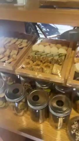 The video shows the display case of a bakery. There are several different varieties of cookies and other baked goods arranged on wooden shelves. The trays and individual portions are covered in clear plastic. There are jars containing more cookies on the bottom shelf. The bakery is well lit.