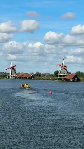 A drone shot captures a wide body of water with a small yellow and black boat in the center. Two windmills stand in the background on the far bank. The sky is blue with scattered white clouds. The windmills are traditional Dutch style, with dark bodies and red and orange sails. The boat is moving away from the camera, leaving a wake behind it. The water is choppy, with small waves. The overall scene is peaceful and scenic, with a distinctly Dutch feel.