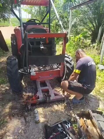 A man is working on the rear of a red tractor, which is supported by a metal frame. He is kneeling and appears to be using tools. There are tools and a spray can on the ground in front of the tractor. The scene is outdoors with trees and vegetation in the background.