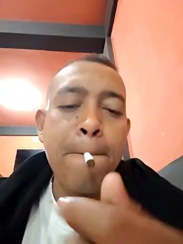 Here is a brief description of the video:
A man with short, dark hair is shown from the chest up, facing the camera. He is holding a cigarette in his mouth and a lighter. He lights the cigarette and exhales smoke toward the camera. The background consists of a pink wall and a dark horizontal bar near the top of the frame.
