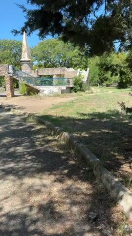 The video pans across an outdoor scene on a sunny day. In the foreground, a gravel path with dappled shade from overhanging trees leads towards a raised, stone structure. This structure appears to be partly ruined, with stone walls, archways, and a conical tower topped with a spire. A blue railing runs along a raised walkway or platform. The surrounding environment consists of green grass, scattered dirt patches, and mature trees under a clear blue sky. The overall impression is of an old, possibly historical, outdoor site.