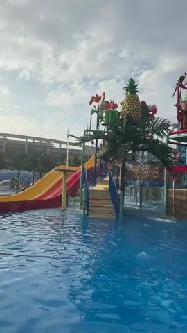 A wide shot of a water park on a cloudy day. The foreground is dominated by a bright blue swimming pool. In the midground, a colorful water play structure with slides, stairs, and various aquatic features stands prominently. A large bucket designed to dump water sits at the top, adorned with decorative parrots. A pineapple decoration is visible on the left side of the structure, along with palm fronds. Water sprays from various points of the structure, creating a lively atmosphere. In the background, a multi-story building is partially visible, suggesting a resort or hotel. The overall impression is of a fun and exciting recreational environment. There are no people clearly visible in the shot.