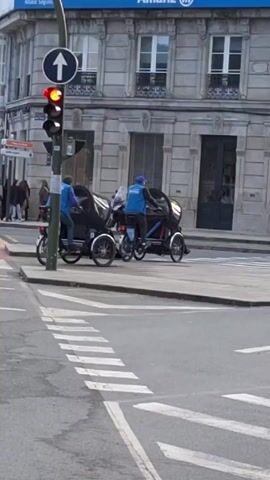 The video shows two men on a bicycle-like carriage waiting at a red light on a city street. Both men are wearing blue vests, and one has a blue hat. The carriage they are on has a black passenger compartment. Behind them, pedestrians cross the street. The building on the corner has an "Allianz Seguros" sign. The traffic light has a red light and a sign indicating traffic is allowed to go straight. The street is marked with a pedestrian crossing. Other cyclists pass by in the background.