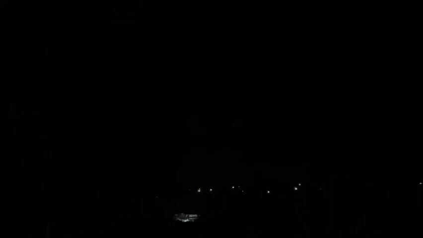 The video displays a static, dark nighttime scene. The foreground and much of the mid-ground are rendered in near-total blackness due to extremely low light conditions. Along the lower third of the frame, a horizontal line of approximately ten small, faint, stationary artificial light sources is visible in the distant background. No specific objects, people, or discernible actions are present.