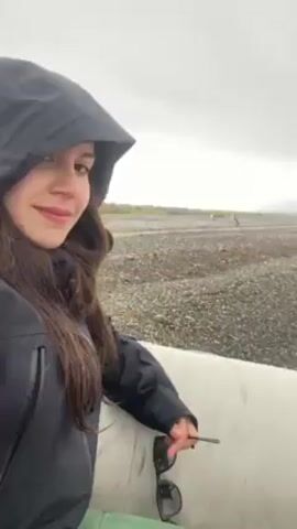 A woman wearing a black hooded jacket and green pants sits inside a white inflatable boat, holding a pair of sunglasses. She smiles towards the camera while the camera pans down and reveals two penguins standing near the edge of the boat on a pebble beach. The sky is overcast and the beach extends into the distance.