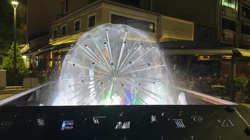 The video captures an illuminated fountain at night in an urban setting, likely in Karditsa, Greece, given the provided context. The central feature is a large, spherical fountain composed of numerous jets of water that spray outwards, creating a dynamic, dome-like shape. Inside the sphere, multi-colored lights cycle through shades of blue, purple, and green, casting vibrant reflections on the water.

In the foreground, a dark, rectangular structure borders the fountain, featuring illuminated, stylized geometric patterns or symbols in blue and purple lights. The background reveals several multi-story buildings, some with lighted windows, suggesting commercial or residential use. To the right, an outdoor seating area of what appears to be a cafe or restaurant is visible, with several patrons seated at tables enjoying the evening ambiance. Streetlights and other artificial lights illuminate the scene. The overall activity centers around the aesthetic display of the fountain and the evening leisure of people in the background. No clear, readable text is discernible on any signs or structures within the video frame. The weather appears calm, and the time of day is clearly night.