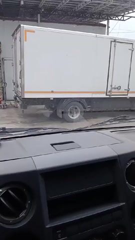 The video is taken from inside a vehicle, looking out the windshield. A white box truck is parked outside a building with a covered loading area. A person in a dark coat walks behind the truck pushing a stack of pallets. The person is not clearly identifiable. The video focuses on the scene outside the vehicle. The interior of the vehicle is partially visible, including the dashboard and air vents. A blue baseball cap is briefly placed on the dashboard.