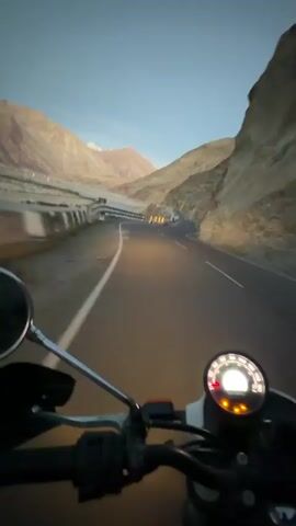 A first-person perspective video shows a motorcycle rider on a paved road through a mountainous desert landscape. The sky is a clear, pale blue, suggesting dawn or dusk. The road curves to the right, with a rocky mountain face on the right and a valley with a river or dry riverbed on the left. The motorcycle's handlebars and dashboard are visible in the foreground, with the speedometer illuminated. The road has white dashed lines and a solid white line on the edge. There are guardrails along the left side of the road and some yellow reflective markers. The mountains in the distance are beige and pink, with snow-capped peaks visible. The overall scene conveys a sense of solitary travel through a vast and rugged environment.