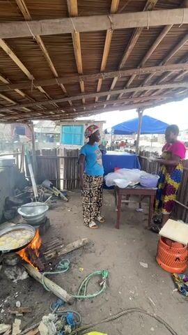 In Orimedu, Nigeria, two women are engaged in a cooking activity under a rustic, open-air shelter. The shelter's roof is constructed from interwoven palm fronds supported by wooden beams, suggesting a practical, possibly temporary structure. The ground is sandy earth, typical of coastal or rural Nigerian environments.

The primary subjects are two women. One woman, on the left, is wearing a blue t-shirt and patterned trousers, with a headscarf. She is actively tending to a cooking fire. The fire is built on the ground, with logs of wood burning intensely. A large metal pot is suspended over the fire, and what appears to be food, possibly fritters or fried dough, is cooking in hot oil within the pot. The woman is holding a long stick or utensil, suggesting she is stirring or managing the food.

The second woman, on the right, is wearing a vibrant, multi-colored traditional dress and a headscarf. She stands near a wooden table covered with a blue cloth. On the table are plastic containers and what look like supplies for food preparation or packaging, including white plastic bags. She appears to be engaged in handing something to or receiving something from the first woman, or perhaps organizing items on the table.

The scene depicts a bustling, informal food preparation area. The environment is somewhat cluttered, with various objects on the ground, including ropes and what looks like discarded materials, indicative of an active and working space. In the background, beyond the immediate cooking area, there are glimpses of other structures, some with blue tarpaulin roofs, hinting at a larger market or community gathering.

The time of day appears to be daytime, with natural light illuminating the scene, though the exact time is not determinable. The weather seems fair, with no indication of rain. The overall atmosphere is one of everyday life and commerce, with the women focused on their tasks. No text is visible in the image.