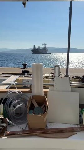This media file depicts a daytime scene at a port in Palamós, Spain. The foreground is filled with various construction or repair materials, including a large roll of clear plastic sheeting, coiled hoses, a cardboard box with sanding discs, and white rectangular boards. These objects suggest ongoing work or preparation for a project.

In the middle ground, a stone pier extends into the water, featuring a large dark bollard. A metal pole with electrical connections and a white control box stands on the pier. The water is a deep blue, reflecting the bright sunlight with a shimmering effect, indicating a sunny day.

The background is dominated by a large cargo ship with a prominent superstructure, anchored or maneuvering in the sea. The ship appears to be a bulk carrier or a similar type of vessel, with cranes and containers visible on its deck. The name "ADRIAN A" is partially visible on the ship's hull. Distant hills or mountains form the horizon line under a clear, bright blue sky, with a few scattered birds flying. The overall mood is one of maritime activity and potentially industrial work within a picturesque coastal setting. The bright sunlight and clear sky suggest fair weather.