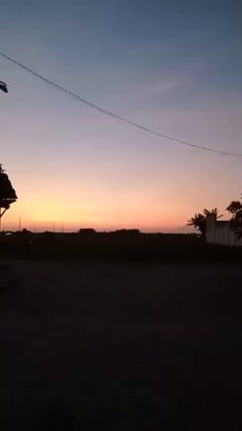 The video captures a serene sunrise or sunset in Bungur, Indonesia. The sky displays a beautiful gradient of colors, transitioning from dark blue at the top to lighter blue, then purplish-pink, and finally a vibrant orange and yellow hue near the horizon.

In the foreground, the ground is dark and unpaved, suggesting a rural or semi-rural setting. To the left, a wooden structure, possibly a small shelter or roadside stall, is silhouetted against the colorful sky. A street lamp on a pole is also visible in silhouette, along with power lines stretching across the upper portion of the frame.

The horizon features a dark silhouette of distant trees or vegetation, and a faint outline of what appears to be a mountain or large hill on the far right. Palm trees and another building-like structure are also visible in silhouette on the right side of the frame.

No people are visible. The only activity is the gradual visual change of the sky, accompanied by the distinct sound of a bird chirping, suggesting it is dawn. The overall atmosphere is peaceful and tranquil.