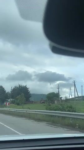 The view from inside a vehicle driving along a road shows overcast skies. The road is divided by a solid white line, and a silver guardrail runs alongside. Road signs indicating a curve in the road are visible. Across the road is a collection of buildings and structures, some partially obscured by trees. There's a structure with a large, flat roof and industrial equipment. Utility poles are also visible in the distance. The vehicle's rearview mirror partially obstructs the view from above.
