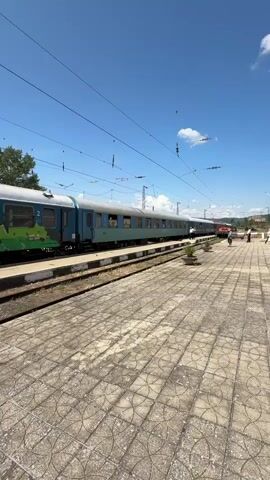 A train sits at a station on a bright sunny day. The train is blue and green, with graffiti on some of the cars. The train is on tracks, and there is a platform next to the tracks. The platform is made of stone tiles and has potted plants on it. There are a few people on the platform. The sky is blue with a few white clouds. There are power lines above the train.