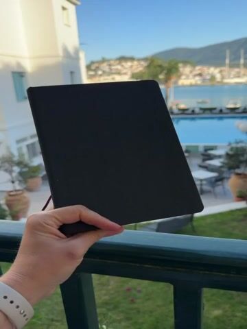 A person's hand, wearing a white smartwatch on the wrist, holds up a black sketchbook against a vibrant outdoor backdrop. The video opens with the closed sketchbook, then quickly transitions to reveal an open spread filled with colorful watercolor sketches.

The left page of the sketchbook features a terracotta-colored artistic rendering of a bust or sculpture with flowing, leaf-like adornments, possibly depicting a mythological figure. Below it, a detailed illustration of a Greek salad is visible, complete with red tomatoes, green cucumber, dark olives, and feta cheese. Greek text, including "Ακροκέραμο" (Akrokeramo), is written above and around the drawings.

The right page displays a watercolor street scene depicting traditional buildings with arched entrances and a warm, pink-hued sky above. Several Greek words are prominently featured: "Ευχαριστώ" (Efcharisto - Thank you), "Καλημέρα" (Kalimera - Good morning), and in large, bold letters, "Πόρος" (Poros). Another word, "Γειά σου" (Gia sou - Hello/Goodbye), is partially visible.

The background shows a quintessential Mediterranean scene, fitting for Poros, Greece. A white building with subtle architectural details is visible on the left. Below, a green railing frames a view of a sparkling blue swimming pool, surrounded by lounge chairs and tables. In the distance, a charming town or city with white and light-colored buildings sprawls along a coastline, backed by rolling hills or mountains, and a clear blue body of water. The sky is bright and clear, suggesting a sunny day.