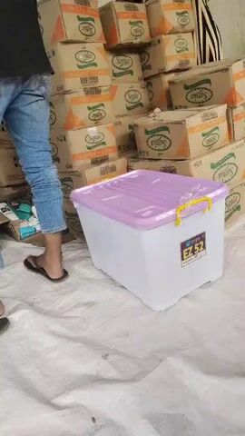 The video shows a person wearing blue jeans and black flip-flops standing in front of a large stack of cardboard boxes. The person is mostly out of frame, but their legs and feet are visible. The boxes are stacked high and appear to contain products. In the foreground, there is a large, clear plastic storage bin with a purple lid. The bin has a label on the side. The floor is covered with a white tarp. The lighting suggests an indoor setting, possibly a warehouse or storage area.