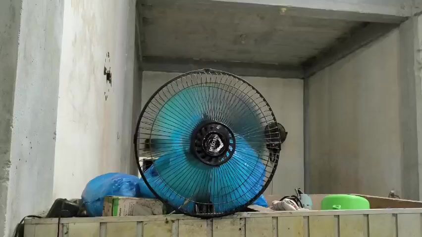 The video shows a dark-colored electric fan with blue blades sitting on a cluttered shelf. The fan blades are rotating, creating a blurred effect. The shelf appears to be constructed from concrete blocks or a similar material, and is lined with light-colored tiles along the top edge. The background consists of unfinished concrete walls and a ceiling. There are various objects on the shelf, including a blue plastic bag, a green container, and other miscellaneous items.