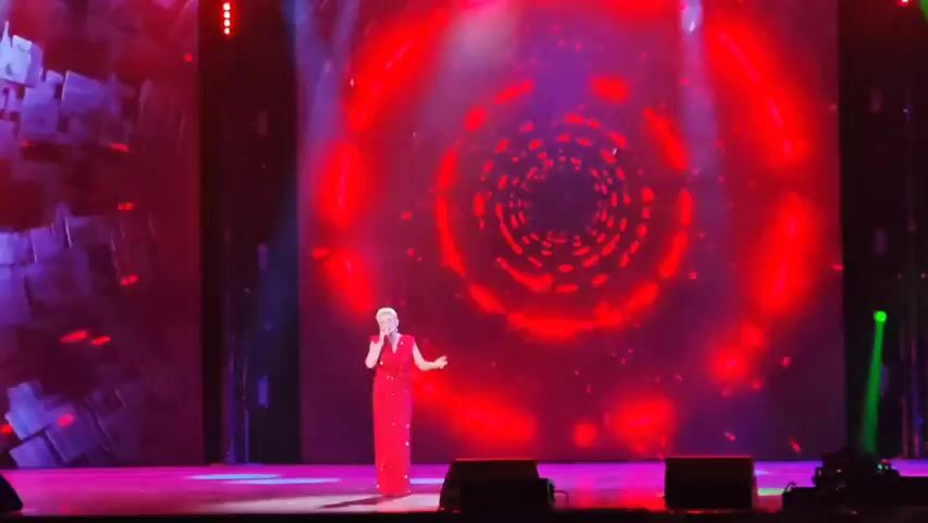 A woman in a red dress stands on a stage holding a microphone. She is singing in front of a large screen displaying a red, swirling, vortex-like image with bright lights. The stage is lit with red and green spotlights.