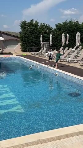 The video shows an outdoor swimming pool area on a bright, sunny day. The sky is clear with a few scattered clouds.

Two young boys are the main subjects. One boy, wearing a white t-shirt and dark shorts, is initially holding a long-handled pool cleaning net. The other boy, wearing a green t-shirt and dark shorts, is standing nearby.

The pool is large and rectangular, with a blue tiled bottom. Numerous light-colored lounge chairs with dark frames and matching umbrellas are neatly arranged along the pool deck, suggesting a resort or hotel setting. Tall, dark green trees form a lush backdrop behind the lounge chairs. In the distance, a building with a red-tiled roof is visible to the left, and a multi-story light-colored building can be seen to the right.

At the beginning of the video, the boy with the net is maneuvering it near the edge of the pool. He then places the net down, and the other boy, who was standing beside him, picks up a soccer ball that was on the pool deck. The boy in the green shirt then throws the soccer ball into the swimming pool, where it floats on the surface. The boys appear to be playing. No visible text is clearly discernible in the video.