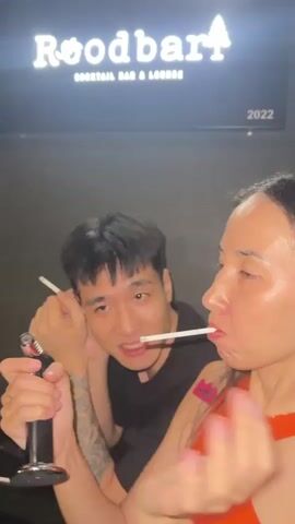 A man with short dark hair and a black t-shirt is in the background, holding a cigarette in his left hand and a lighter in his right hand. He is looking towards the right. In the foreground, a woman with long dark hair and an orange top is holding a cigarette in her mouth. The man is using the lighter to ignite the cigarette in the woman's mouth. Smoke is visible. A neon sign with the word "Roodbar" and a tree graphic is visible in the background, along with the year "2022".