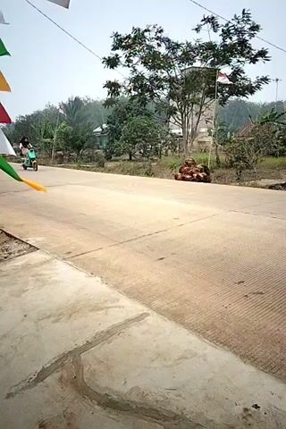 A person rides a light blue and yellow electric scooter on a paved road. They are wearing a hijab and a light-colored outfit. The scooter is moving from left to right in the frame. The road is in the foreground, with a concrete curb on the left. In the background, there are houses, trees, and Indonesian flags on poles. The sky is overcast. The person and the scooter are the main subjects of the video. The video appears to be shot outdoors in a rural or suburban setting.
