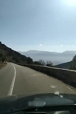 A car is driving on an asphalt road with a white line in the center. The road winds through a mountainous, wooded area.  To the left, there is a mountainside covered in trees and shrubs. To the right, there is a low stone wall along the edge of the road. In the distance, mountain ranges are visible, appearing hazy under a bright, cloudless, blue sky. The sun is shining brightly, causing lens flare in the camera. The view is from inside the car, looking out over the dashboard.