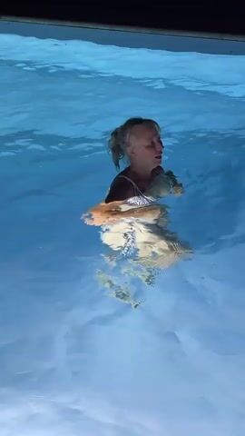 The video begins with a woman swimming in a pool. The water is a bright blue and reflects the sunlight. The woman is wearing a white bikini and has her hair tied back. She appears to be enjoying herself, smiling and looking around the pool.

The scene then transitions to the same woman sitting on the edge of the pool, with her feet in the water. She is wearing a patterned dress. The lighting suggests it is evening or nighttime, as the sky is dark and there are lights illuminating the pool, creating a blue glow. She is smiling and appears relaxed.

The setting is a swimming pool, likely outdoors, given the presence of the night sky and surrounding vegetation visible in the background. The time of day is evening or night. There is no visible text in the video. The location is Saint-Galmier, France. The overall mood is one of leisure and enjoyment of the swimming pool.