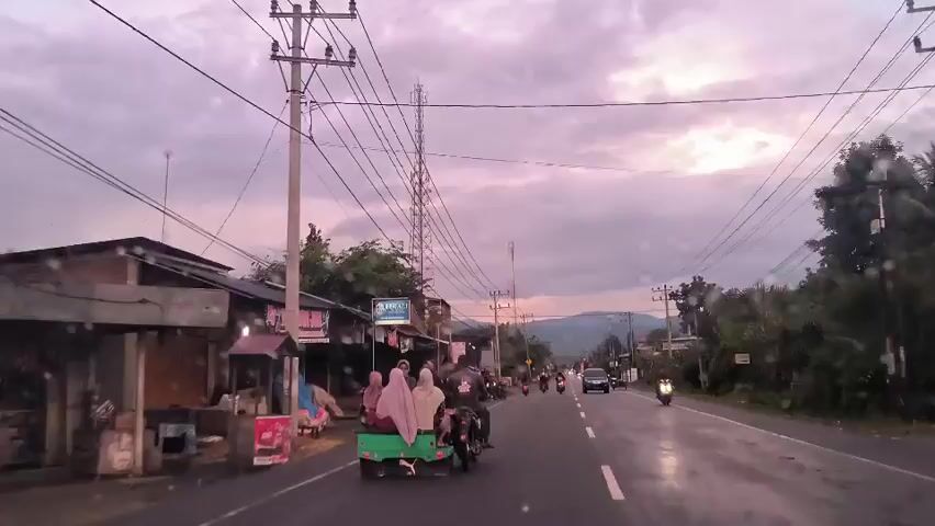 The video shows a car driving on a paved road in a rural area. On the side of the road are small buildings, some of which appear to be shops. There are power lines running along the road. Several people are traveling on motorcycles. One motorcycle has a small trailer attached, in which two women wearing headscarves are riding. In the distance, there are mountains. The sky is overcast. There are raindrops on the windshield of the car.