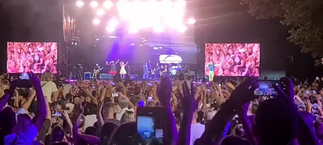 The video captures a live concert with a band performing on a brightly lit stage. Two large screens display live footage of the performers. A large, enthusiastic crowd is visible in the foreground, with many people holding up their phones to record or photograph the event. The audience is packed together, and many arms are raised in the air. The stage is set up with musical instruments, including drums, guitars, and microphones. The lighting is dynamic, with bright spotlights and purple hues illuminating the stage and parts of the crowd. The overall atmosphere appears to be energetic and celebratory.