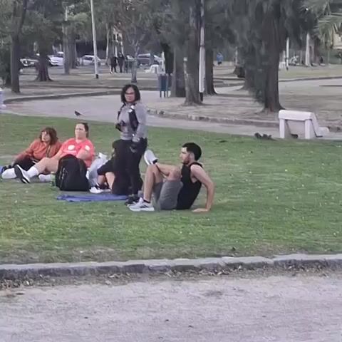 The video captures an outdoor scene in a park in Villa Domínico, Argentina, during the daytime. The lighting suggests diffused sunlight, possibly from an overcast sky or late afternoon.

In the foreground, a group of five people is gathered on a grassy area. A man wearing a black tank top, grey shorts, and white and grey athletic shoes is seated on the grass, leaning back on his hands. He is performing a leg stretch, with one leg bent and his foot resting on the knee of his other leg.

Standing nearby is a woman in a grey long-sleeved top and black pants. Behind the man and the standing woman, two women are seated on a blue mat or blanket on the grass. One woman wears a reddish-orange top, and the other wears a black top. A black backpack is visible next to them. A fifth person is partially obscured behind the woman in the black top.

The setting is a typical park environment, featuring expansive green lawns, paved walking paths, and numerous tall trees, including some dense evergreen-like trees and a palm tree. White park benches are scattered in the background. Other individuals can be seen in the distance, walking along the paths, indicating public use of the park. The overall mood is relaxed, with people enjoying the outdoor space. There is no visible text in the video.